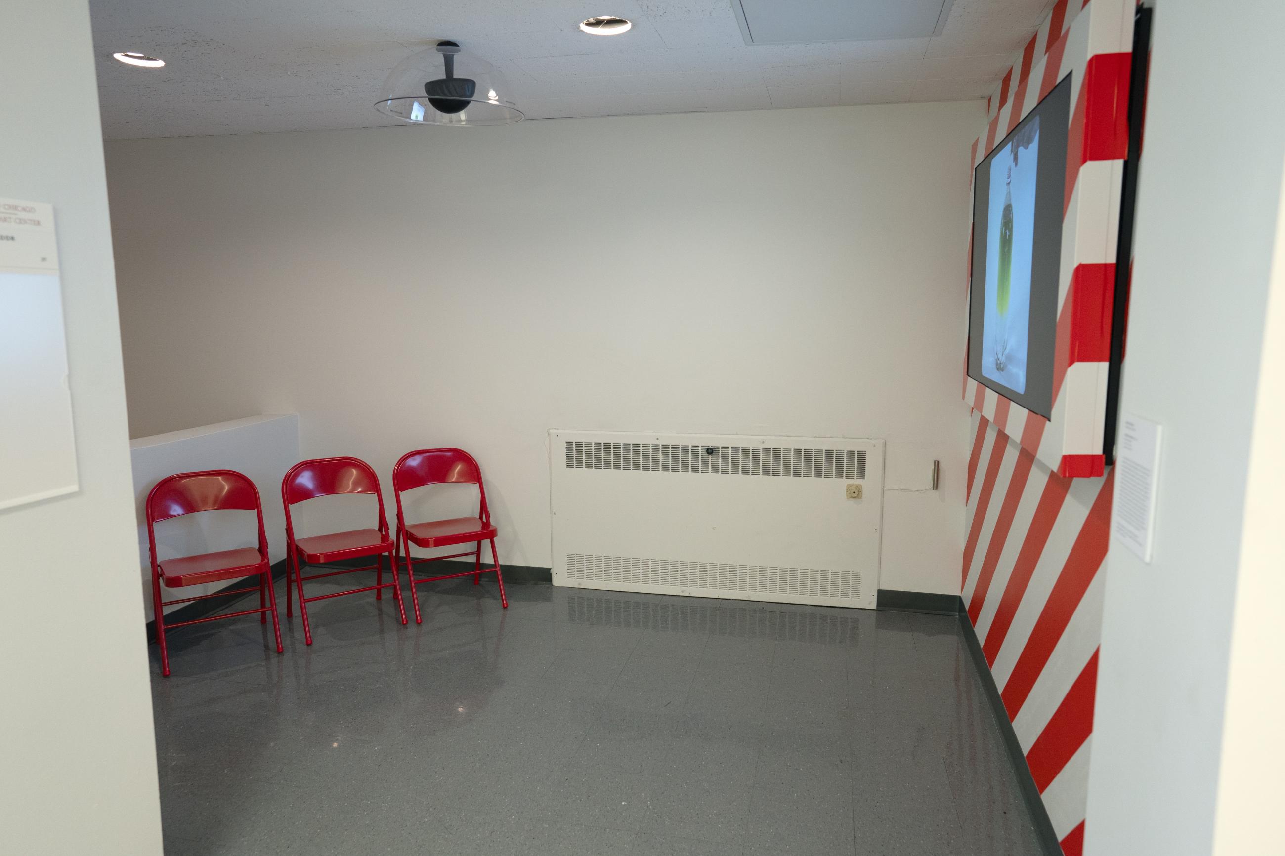 Three red folding chairs in front of a monitor on a red and white striped wall