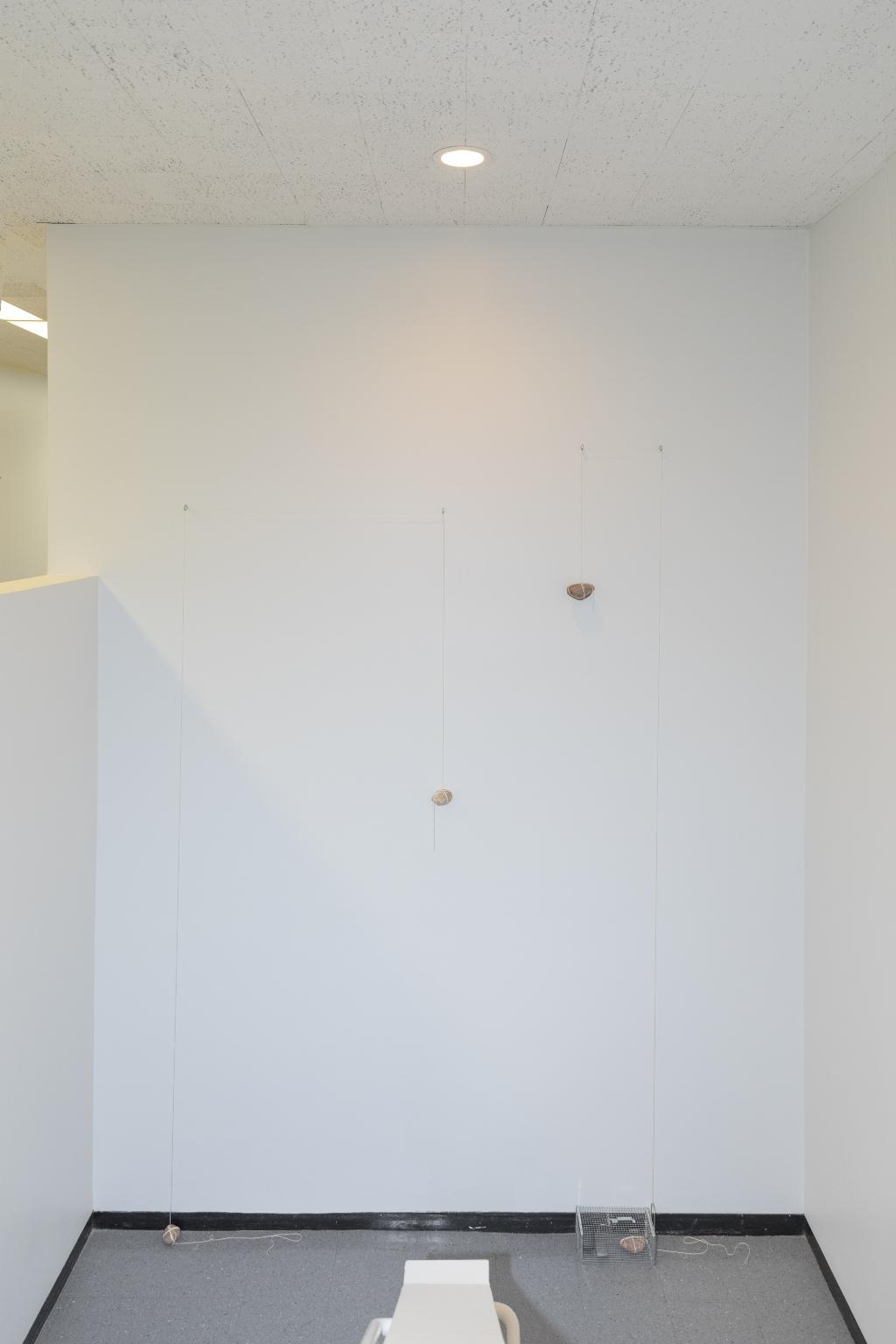 Two rocks hang from strings on teh wall, while two other rocks anchor the work to the floor. One of the rocks on the floor is in a cage.