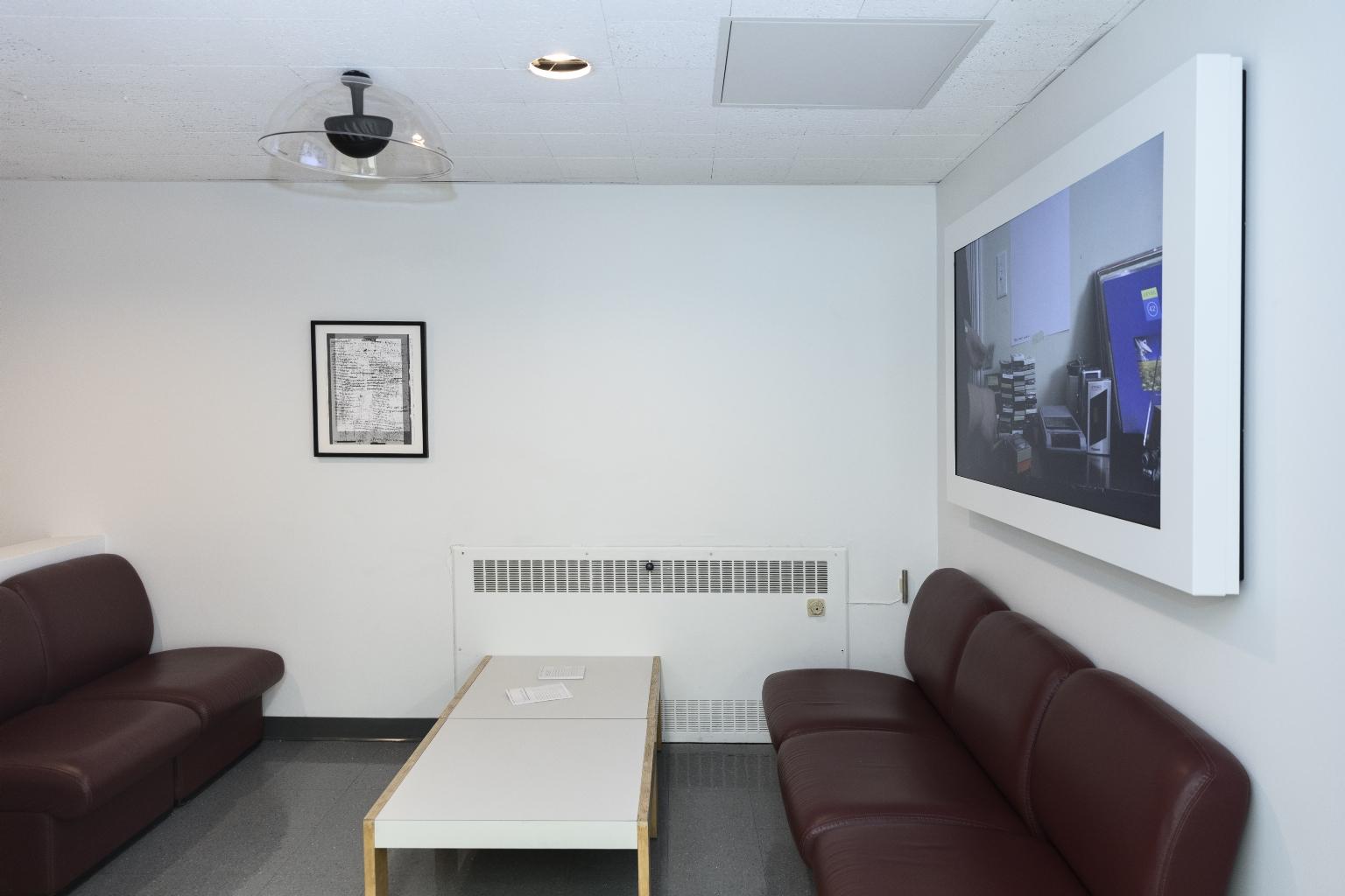 View of framed artwork and video artwork in seating area with cushioned chairs and a table.