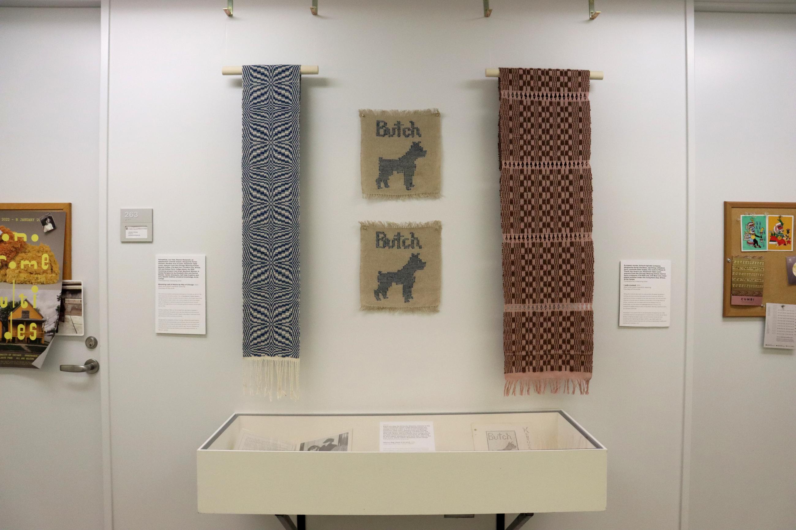 A display of woven textiles hanging on a white wall above a display case