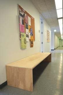 long wooden bench in a white hallway with with bulletin board covered in posters above it